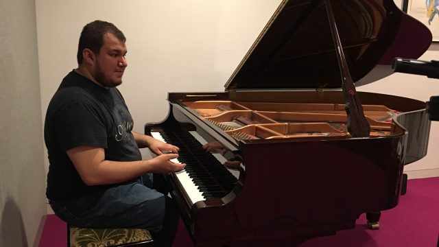 Hector sits at a grand piano before a camera and microphones during a recording session.
